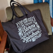 Load image into Gallery viewer, black tote bag with white text on a chair in a room setting