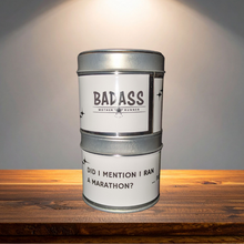 Load image into Gallery viewer, Candle inside a tin with 'BADASS' label on a wooden surface