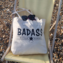 Load image into Gallery viewer, Tote bag with 'BADASS' text on a chair by a pebbly beach

