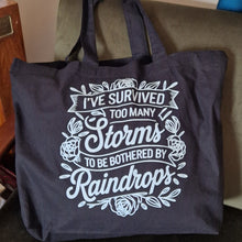 Load image into Gallery viewer, Black tote bag with white text on a green chair
